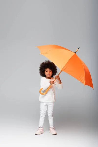 Повний вигляд афроамериканської дівчини, що стоїть з помаранчевою парасолькою на сірому — стокове фото