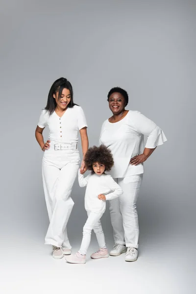 Lächelnde afrikanisch-amerikanische Frauen und Mädchen in weißen Kleidern posieren mit den Händen auf den Hüften auf grau — Stockfoto
