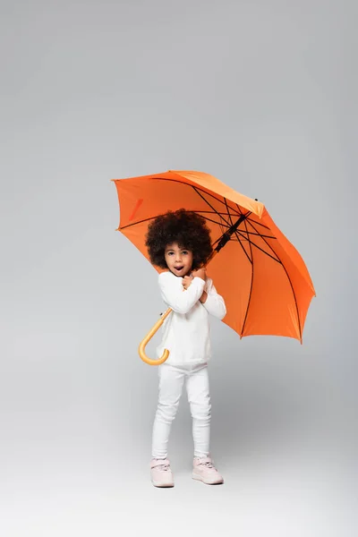 Vue pleine longueur de fille afro-américaine avec parapluie orange debout dans des vêtements blancs sur gris — Photo de stock