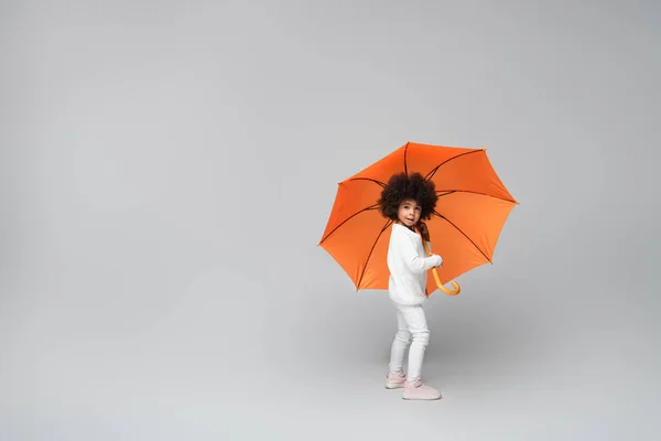 Full length view of stylish african american girl under orange umbrella looking at camera on grey — Stock Photo