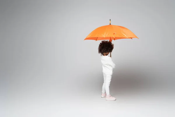 Enfant afro-américain en chemise blanche à manches longues et pantalon debout sous un parapluie orange sur fond gris — Photo de stock