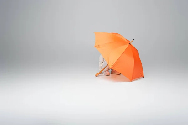 Дитина в білому одязі ховається під помаранчевою парасолькою на сірому фоні з копіювальним простором — стокове фото
