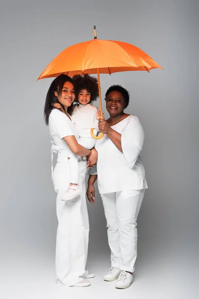 Alegre africano americano mujeres y niño mirando cámara bajo naranja paraguas en gris - foto de stock