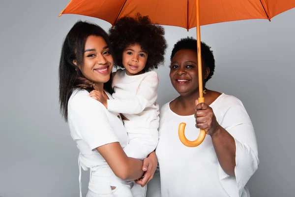 Glückliche afrikanisch-amerikanische Frauen und Mädchen in weißen Kleidern schauen in die Kamera unter orangefarbenem Regenschirm isoliert auf grau — Stockfoto