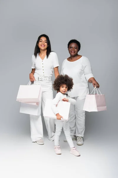 Smiling african american kid with mother and granny holding white shopping bags on grey — Stock Photo