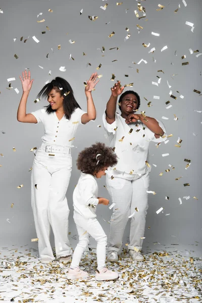Happy african american women and child in white clothes under shiny confetti on grey — Stock Photo