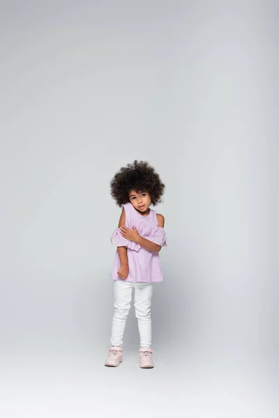 Повнометражний вид на афроамериканську дитину у фіолетовій блузці та білих штанах, що стоять на сірому — стокове фото