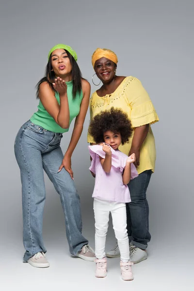 African american woman blowing air kiss near mature mom and daughter on grey — Stock Photo