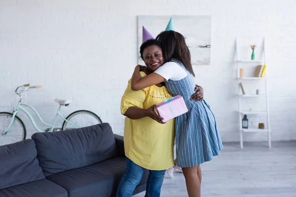 Jovem afro-americano mulher no tampão do partido abraçando feliz mãe segurando presente de aniversário — Fotografia de Stock
