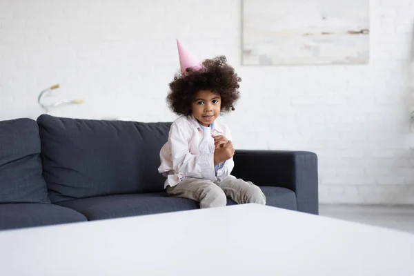 Afrikanisches amerikanisches Geburtstagskind in Partymütze sitzt auf Couch neben verschwommenem Tisch und blickt in die Kamera — Stockfoto