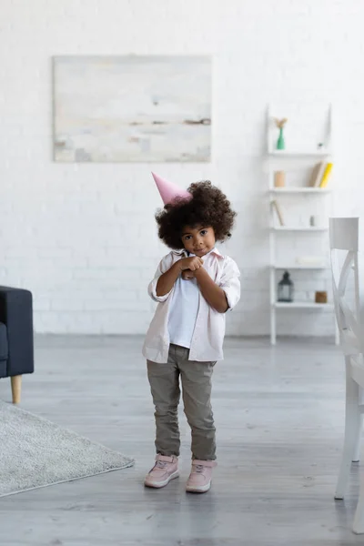 Pleine longueur vue de timide afro-américaine fille en chapeau de fête en regardant la caméra dans le salon — Photo de stock