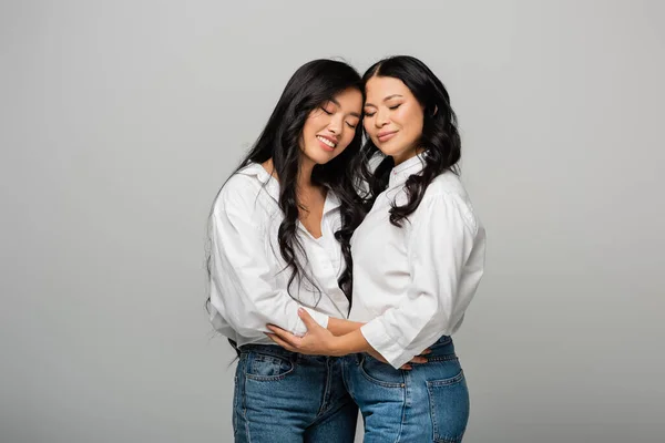 Happy asian mother and daughter in blue jeans and white shirts hugging isolated on grey — Stock Photo