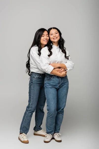 Pleine longueur de heureux asiatique mère et fille en jeans bleus et chemises blanches câlins sur gris — Photo de stock