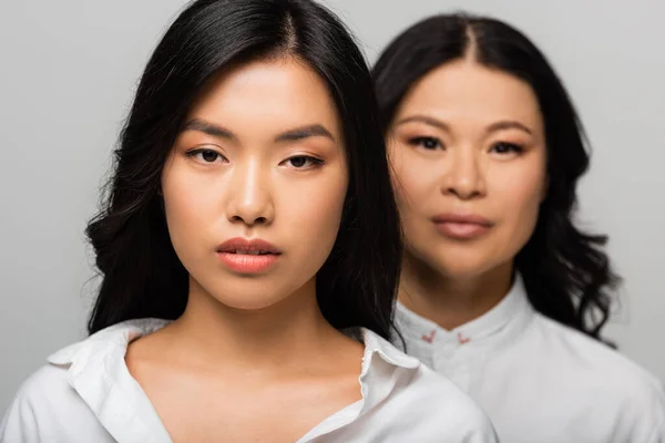 Asian young adult daughter near blurred mother standing behind isolated on grey — Stock Photo