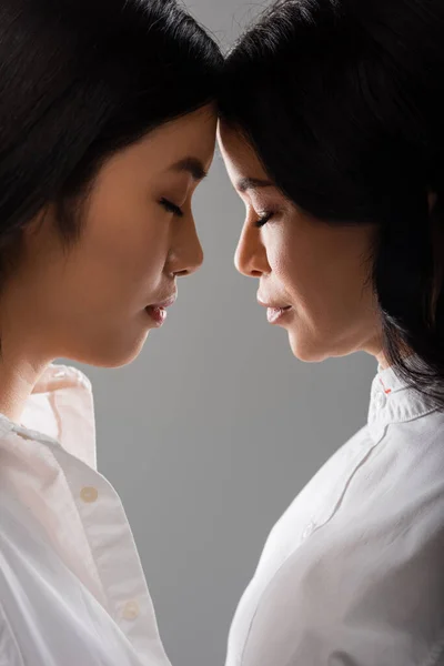 Side view of asian mother with closed eyes leaning on young adult daughter isolated on grey — Stock Photo