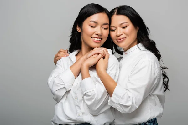 Cheerful asian mother and young adult daughter hugging each other and holding hands isolated on grey — Stock Photo