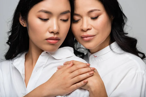 Азіатська брюнетка мати і молода дочка обіймає один одного і тримає руки ізольовані на сірому — стокове фото