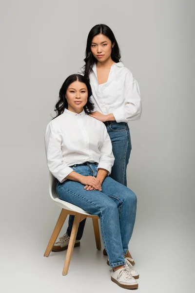Comprimento total da mãe asiática sentada na cadeira perto da filha em jeans azul e camisa branca no cinza — Fotografia de Stock