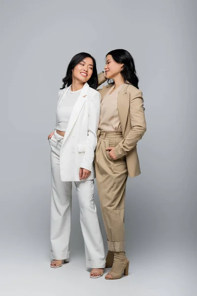 Full length of stylish asian mother and young adult daughter in suits smiling on grey — Stock Photo