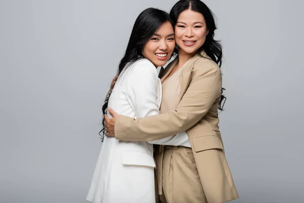 Весела азіатська мати і щаслива молода доросла дочка в костюмі обіймається ізольовано на сірому — стокове фото