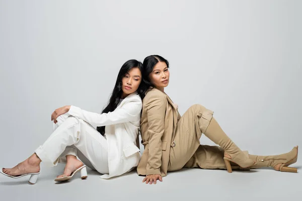 Full length of stylish asian mother in beige suit and trendy young adult daughter sitting on grey — Stock Photo