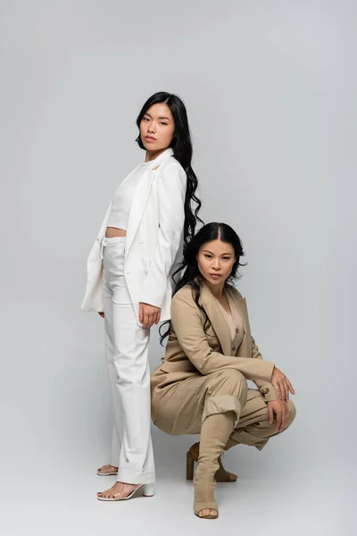 Full length of trendy asian mother in beige suit and stylish young adult daughter posing on grey — Stock Photo