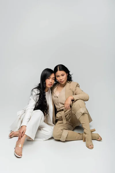 Full length of stylish asian mother in beige suit and brunette young adult daughter sitting on grey — Stock Photo