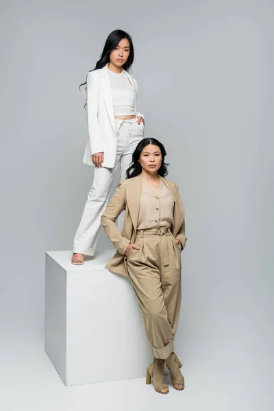 Full length of asian young adult woman standing on cube near brunette mother posing on grey — Stock Photo