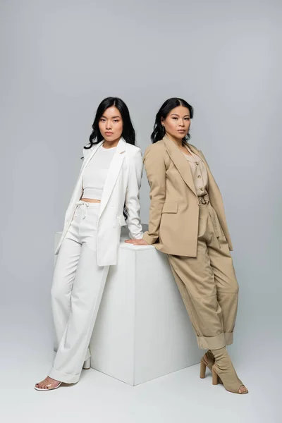 Full length of asian young adult woman standing with brunette mother near cube on grey — Stock Photo