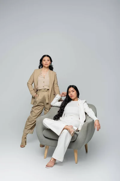 Full length of young adult asian woman sitting in armchair near stylish mother on grey — Stock Photo