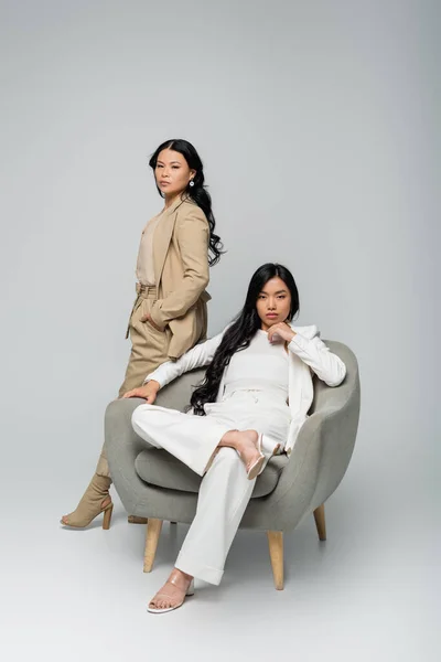 Piena lunghezza di giovane adulto asiatico donna seduta in poltrona vicino trendy madre su grigio — Foto stock