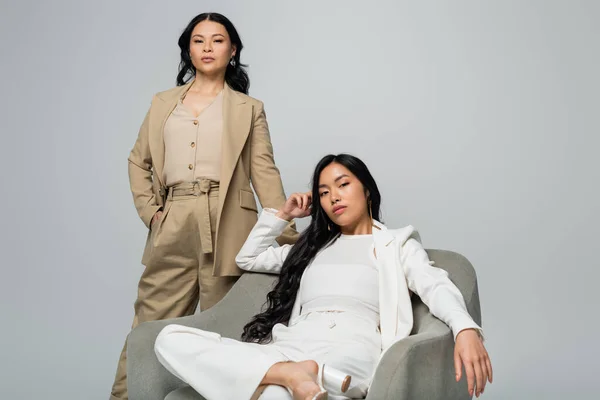 Young adult asian woman sitting in armchair near trendy mother isolated on grey — Stock Photo