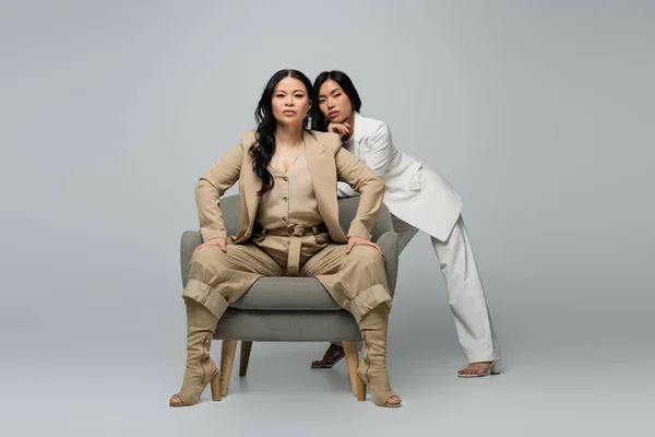 Full length of asian woman sitting in armchair near trendy young daughter on grey — Stock Photo