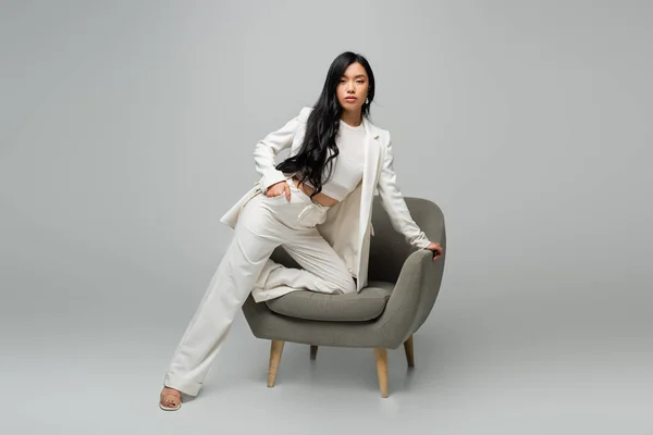Full length of young asian woman leaning on armchair while posing with hand in pocket on grey — Stock Photo