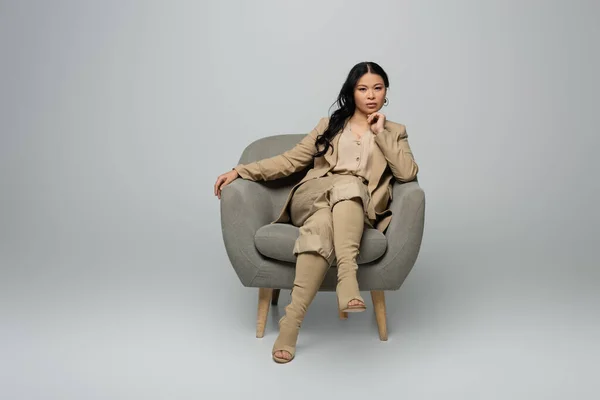 Full length of stylish asian woman sitting in armchair on grey — Stock Photo