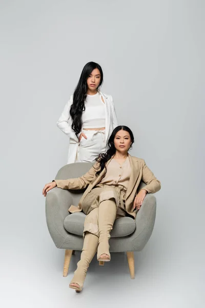 Full length of asian woman sitting in armchair near trendy young daughter standing with hand in pocket on grey — Stock Photo