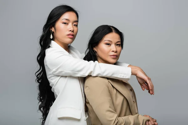 Young asian woman hugging brunette mother in beige suit isolated on gray — Stock Photo