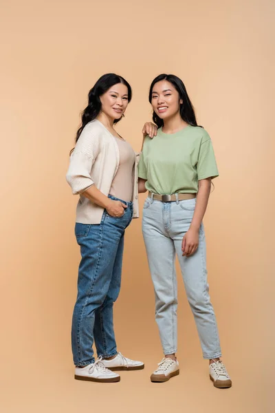 Comprimento total de feliz mãe asiática em jeans de pé com a mão no bolso perto da filha no bege — Fotografia de Stock