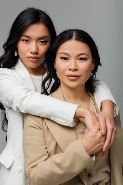 Junge asiatische Tochter umarmt brünette Mutter in beige Anzug isoliert auf grau — Stockfoto