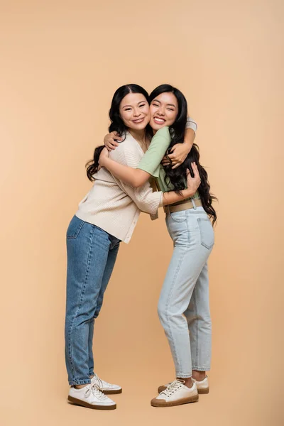 Full length of cheerful asian mother and daughter hugging on beige — Stock Photo