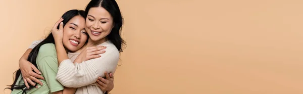 Cheerful asian mother and daughter hugging isolated on beige, banner — Stock Photo