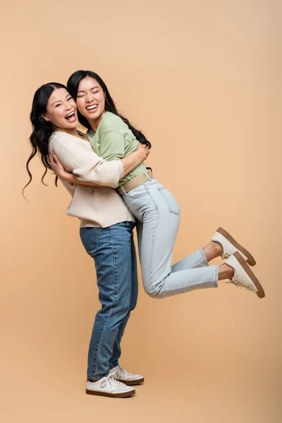 Full length of cheerful asian mother lifting daughter on beige — Stock Photo