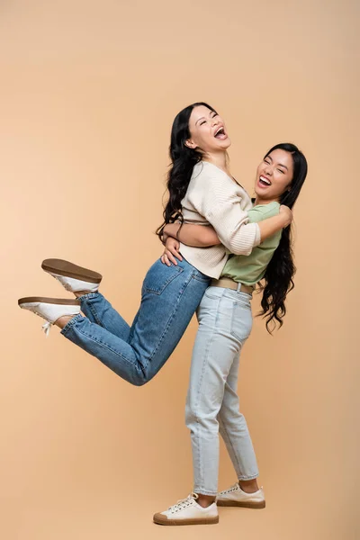 Full length of happy asian daughter lifting excited mother on beige — Stock Photo