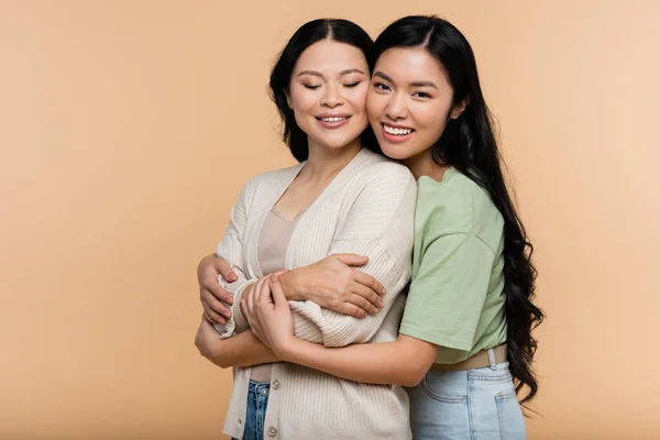 Asian young adult daughter hugging pleased mother isolated on beige — Stock Photo