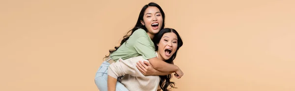 Amazed asian mother piggybacking adult daughter with open mouth isolated on beige, banner — Stock Photo