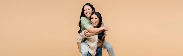 Eccitato asiatico madre piggybacking felice giovane adulto figlia isolato su beige, striscione — Foto stock