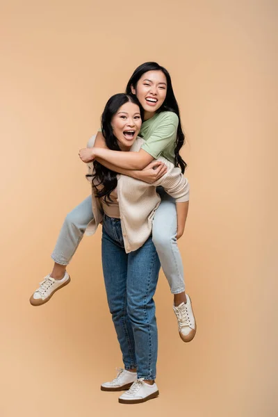 Full length of amazed asian mother piggybacking adult daughter with open mouth on beige — Stock Photo