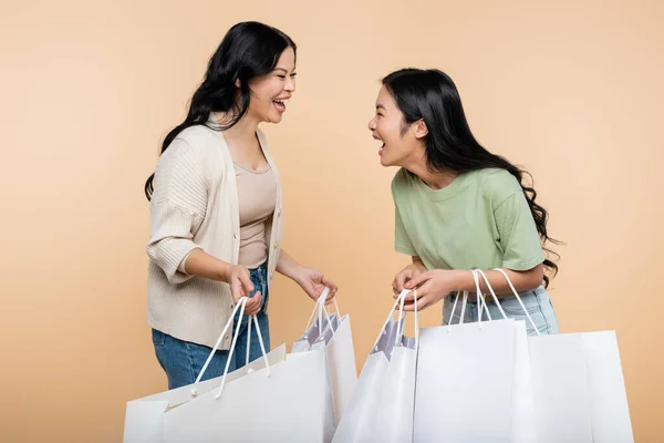 Cheerful asian mother and daughter holding shopping bags and laughing isolated on beige — Stock Photo