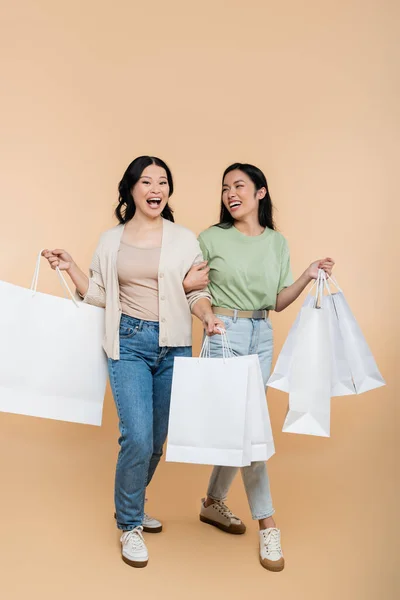 Volle Länge der asiatischen Mutter und junge erwachsene Tochter lächelnd beim Gehen mit Einkaufstaschen auf beige — Stockfoto