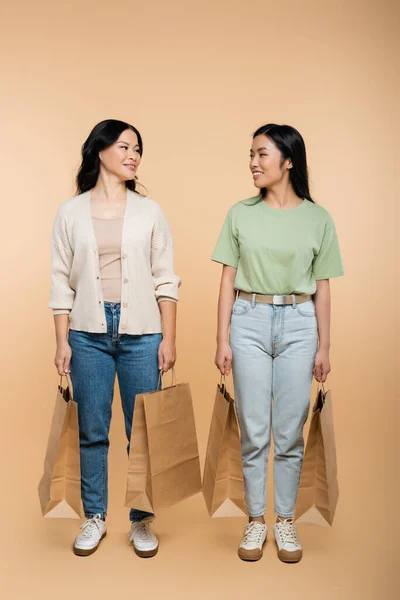 Full length of cheerful asian mother and daughter with paper bags on beige — Stock Photo